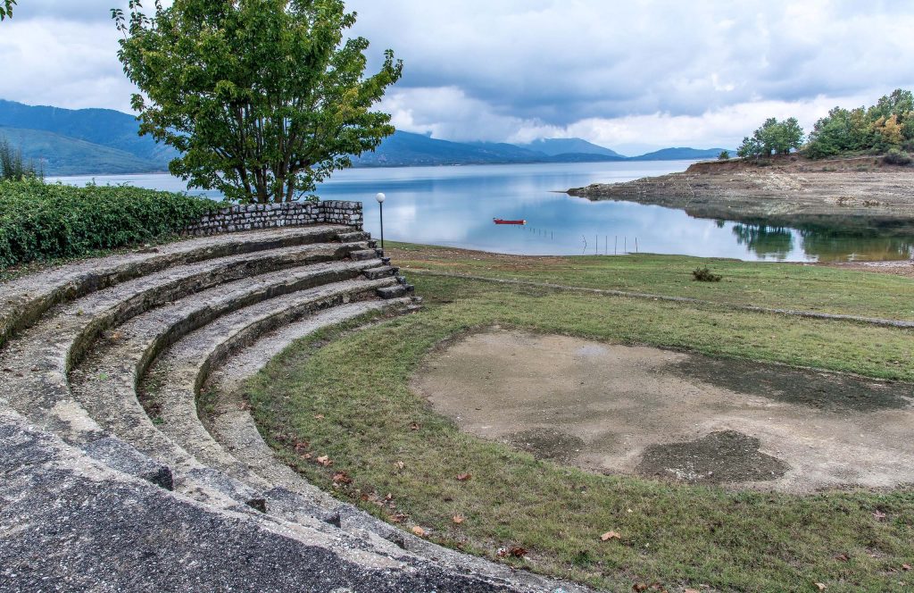 Curved stone steps of a small open-air theatre beside a calm lake, with a red boat on the water and hills behind