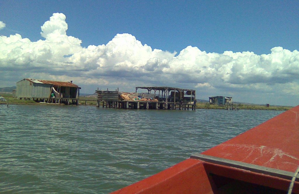 Wooden huts on stilts over the water, seen from a red boat, under large white clouds