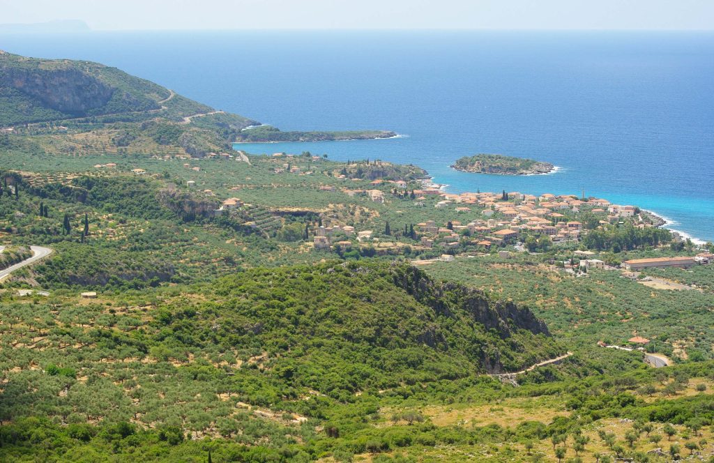 View from a hill over olive groves and a village on the coast, with a small island in the blue sea