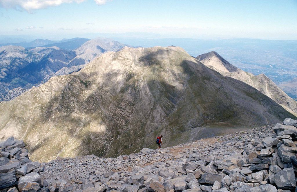 Bare rocky ridge and peaks seen from a summit covered with loose stones, with lowlands far below