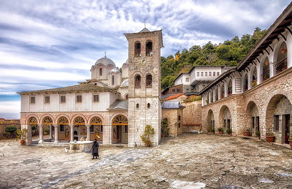 Stone courtyard of Panagia Eikosifoinissa Monastery, with a bell tower and surrounding buildings on a wooded hillside.